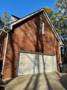 A red brick house with a large white double garage door. The driveway is concrete, and there are tall trees in the background casting shadows on the building. One trash bin is placed beside the garage. The sky is clear and blue.