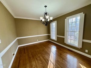 A dining room with hardwood floors, tan walls, and white trim. Two large windows with white shutters let in natural light. A black chandelier with multiple bulbs hangs from the ceiling. The room is empty of furniture.