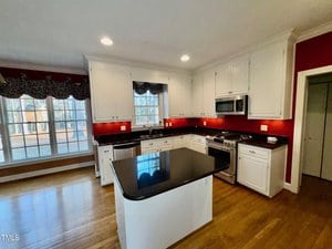 A modern kitchen with white cabinets, stainless steel appliances, and a black granite countertop island. The walls are painted red, and there are large windows with patterned valances. The floor is wooden, and there is recessed lighting.