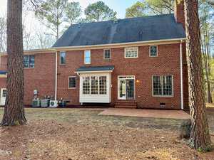 A two-story brick house with a black roof. It has several windows on both floors and a small back door leading to a brick patio. Tall trees surround the house, and the ground is covered with dry pine needles.