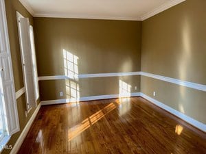 An empty room with green walls and white trim. Sunlight streams through a window, casting shadows on the hardwood floor. The room features crown molding and a white chair rail, with a closed shutter on the left.