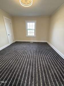 A small empty room with beige walls and a striped gray carpet. There is a single window in the center of the far wall, allowing natural light. A white door is on the left wall. The ceiling features a round, modern light fixture.