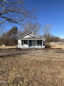 A small, white single-story house with a green front door and two windows. It sits on a grassy lot with sparse trees. A clear blue sky is overhead. A fence is visible on the right side of the property.