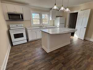 A kitchen with wooden flooring features a central island with a marble countertop. There is a white stove and oven, a microwave above it, a refrigerator, and cabinets. Three pendant lights hang above the island. Two windows are present above the sink.