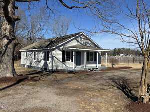 A small, single-story house with light gray siding and a gable roof is surrounded by leafless trees. It features a covered front porch and is set on a sparse lawn with a gravel driveway. The sky is clear and blue.