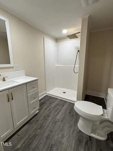 A bathroom featuring a white vanity with a sink and gold handles, a large mirror, a walk-in shower with a rainfall showerhead, and a toilet. The flooring is dark wood, and the walls are painted a light beige color.