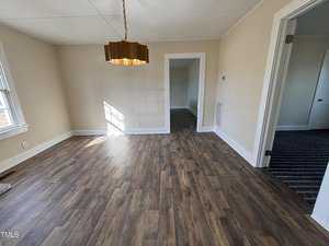 A bright, empty room with light brown wooden flooring and cream-colored walls. A large, geometric pendant light hangs from the ceiling. Sunlight streams through a window on the left, and adjacent rooms are visible through open doorways.
