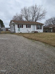 A small white house with a brown shingled roof and maroon shutters sits on a grassy yard. There is a gravel driveway in the foreground, and bare trees are visible in the background. A garden hose is coiled on the side of the house.
