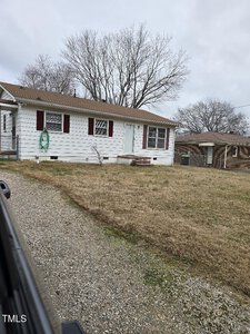 A small, single-story house with white siding and brown shutters is pictured. It has a gravel driveway and a hose coiled on the exterior wall. The yard is dry with patches of grass, and bare trees are visible in the background.
