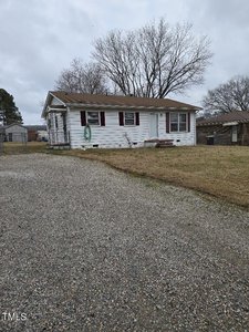 A small white house with red shutters sits on 1851 S Clearview Drive in Henderson, surrounded by a yard with patchy grass. A gravel driveway leads to the home, and a garden hose hangs on the side. Leafless trees rise in the background under a cloudy sky.