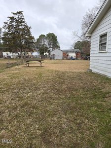 A grassy backyard with a wooden picnic table in the middle. A white house is visible on the right side, with a storage shed and trees in the background. The sky is overcast.