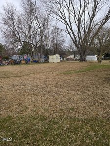 A large backyard with brown grass, bordered by leafless trees and a wooden fence. Several sheds and scattered items are visible at the back. The sky is overcast, suggesting a cloudy day.