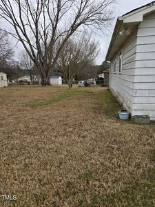 Side view of a white house with a grassy yard. A large leafless tree is near the house. In the background, there are more trees and a small shed. The sky appears overcast.