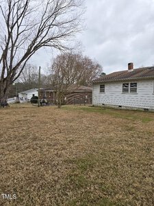 A grassy backyard with a large tree on the left and two single-story houses in view. The sky is overcast. One house has white siding, and the other is brick. A wooden fence runs between the houses.