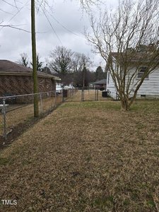 A narrow backyard with a chain-link fence on the left. The grass is brown, and there are bare trees. A white house is partially visible on the right. Overcast sky in the background.