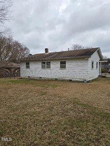 A small, single-story white house with a sloped roof and chimney is situated on a grassy lawn. The house has several windows and appears weathered. The sky is overcast, and there are bare trees surrounding the property.