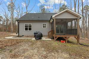 A small house with gray siding and a black roof is surrounded by a wooded area. The back features a screened porch and a grill. The yard is covered with dry leaves and patches of grass. The sky is partly cloudy.