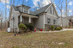 A two-story house with gray siding and stone accents is surrounded by leafless trees. It has a covered porch, multiple windows, and a dark roof. Three red planters are placed beside the staircase leading to the front door. The sky is partly cloudy.