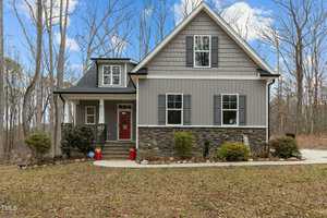 Nestled in a wooded area, the two-story house at 2482 Golden Forest Drive in Franklinton boasts gray siding, stone accents, and white trim. A charming front porch with white railings and a red door invites visitors up the steps, flanked by trees and a convenient driveway to the right.