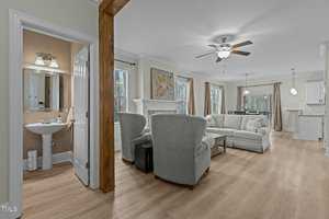 Spacious living area with light wood flooring, featuring a beige sofa and two armchairs facing a fireplace. A ceiling fan and recessed lighting illuminate the room. An open doorway leads to a small, neatly arranged bathroom.