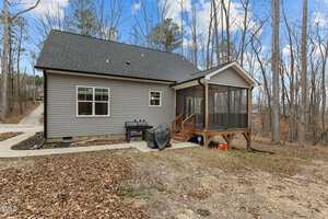 A small house with gray siding and a dark roof is situated in a wooded area. It features a screened porch with wooden steps. A black grill and a covered item are near the porch. The ground is covered with dry leaves. A concrete path leads to the house.