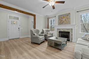 A living room with light wood flooring, featuring a ceiling fan. It has two armchairs and a sofa, a large coffee table, and a fireplace with artwork above it. Two windows on either side of the fireplace allow natural light into the room.