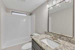 Bathroom with a granite countertop and a sink. Above the sink is a large mirror with three light fixtures. To the left is a white bathtub with tiled walls and a small window. The walls are light-colored.