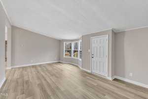 A spacious living room with light wood flooring, gray walls, and a white ceiling. The room features a bay window on the right with a view of trees and a white door near the window, leading outside. The space is empty and well-lit.