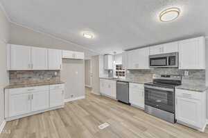 A modern kitchen with light wood flooring and white cabinets. It features a stainless steel stove, microwave, and dishwasher. The countertops are granite with a gray tile backsplash. Ceiling lights illuminate the space.