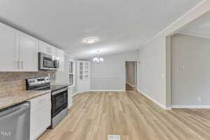 A modern kitchen and dining area with wooden flooring. The kitchen features white cabinets, stainless steel appliances, and a brick-patterned backsplash. There's a ceiling light fixture, and an open dining space with a small built-in cabinet.