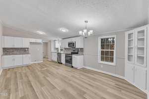 A modern kitchen and dining area with white cabinets, stainless steel appliances, and a light wood floor. There's a window on the right, letting in natural light, and a chandelier hanging above. A small built-in corner cabinet is visible on the right wall.