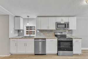 A modern kitchen with white cabinets, stainless steel appliances, and a tile backsplash. There is a window above a double sink, a microwave above the stove, and a dishwasher next to it. The floor appears to be wooden.