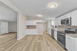 A modern kitchen with white cabinets, stainless steel appliances, and a gray tile backsplash. The room has light wood flooring and a ceiling light fixture. An open doorway leads to an adjacent room with matching flooring and a closed white door.