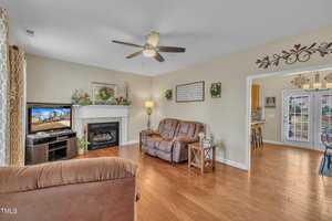 A living room with a brown sofa and recliner set, a TV on a stand next to a fireplace, and a ceiling fan. The room features wood flooring and decorative wall hangings. An open doorway leads to a kitchen area with a glass door to the outside.