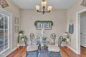 A dining area features a round glass table with four chairs on a blue rug. A sign reading "GATHER" is mounted on the wall, surrounded by greenery. A modern chandelier hangs above. Various plants and decorative items are placed around the room.