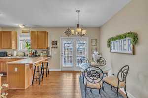Kitchen and dining area with wooden flooring. A kitchen island with stools, wooden cabinets, and appliances is on the left. A dining table with chairs is on the right. French doors lead outside, with a sign reading "GATHER" on the adjacent wall.
