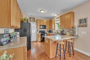 A modern kitchen with wooden cabinets and a black refrigerator. The countertop has two barstools, a toaster, a coffee maker, and various kitchen items. A plant and decorative items are on the cabinets. The floor is wood, and walls are beige.