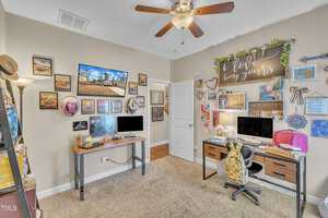 A home office with two wooden desks, each holding a computer. Walls are adorned with various pictures and decor, including a large sign reading "love bring yours." A ceiling fan is above, and a closed white door is in the background. Carpeting covers the floor.