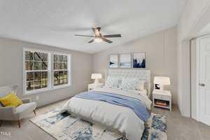 A bedroom featuring a bed with white and blue bedding, two nightstands with lamps, a ceiling fan, a patterned rug, and a gray armchair. A large window offers a view of trees, and there are three abstract paintings on the wall.