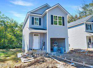 A two-story gray house under construction with white trim and black shutters. The front yard features scattered construction materials. A garage door is open, and a wooded area surrounds the property. The sky is clear and sunny.