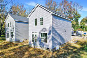 Two newly constructed, identical two-story houses with gray siding and white trim are situated side by side. They have multiple large windows and are surrounded by a grassy area with trees. A vehicle and some construction materials are visible nearby.