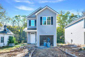 A two-story gray house at 102 Woodson Street is under construction, featuring a peaked roof and single-car garage. The unlandscaped front yard has construction materials scattered about, while trees sway in the background under Oxford's clear blue sky.