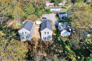 Aerial view of a residential area featuring two gray two-story houses under construction with dark roofs. Surrounding the houses are trees and other homes. The ground appears muddy, and a white truck is parked nearby on a driveway.