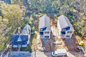 Aerial view of a residential street with three houses. Two modern homes are under construction with visible materials and a parked white truck. The third house is older, with a gabled roof, and surrounded by trees and grass.