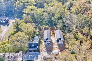 Aerial view of two identical two-story houses under construction, positioned side by side on a lot surrounded by trees. A finished house is nearby, and a road runs in front of the properties. Construction vehicles are visible on site.