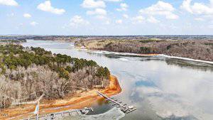 Aerial view of a lake with a mix of calm water and thin ice patches. The shoreline is lined with leafless trees on the right and pine trees on the left. A series of docks extend from the shore. The sky is partly cloudy.