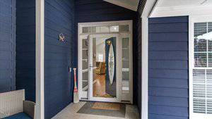 A porch with dark blue siding features a glass-paneled front door with an oval decorative element. A welcome mat lies in front of the door. Indoor furniture is partially visible through the door. A wicker chair and a canoe paddle are on the porch.