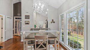 Dining area with a wooden table and four chairs on a rug. Large window on the right with a view of outdoors. Modern chandelier overhead. Light-colored walls and hardwood floor. White doors in the background with small framed pictures.