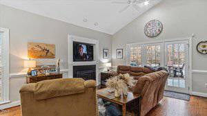 A cozy living room with light gray walls featuring a flat-screen TV above a fireplace, surrounded by brown sofas and chairs. The room has a large window and double glass doors leading to a deck. Various small decorative items decorate the space.