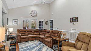 Living room with a large brown sectional sofa and a matching recliner. A blue and white rug covers the floor. Walls are decorated with framed pictures. A round clock is mounted above a set of glass doors leading outside. Ceiling fan is visible above.